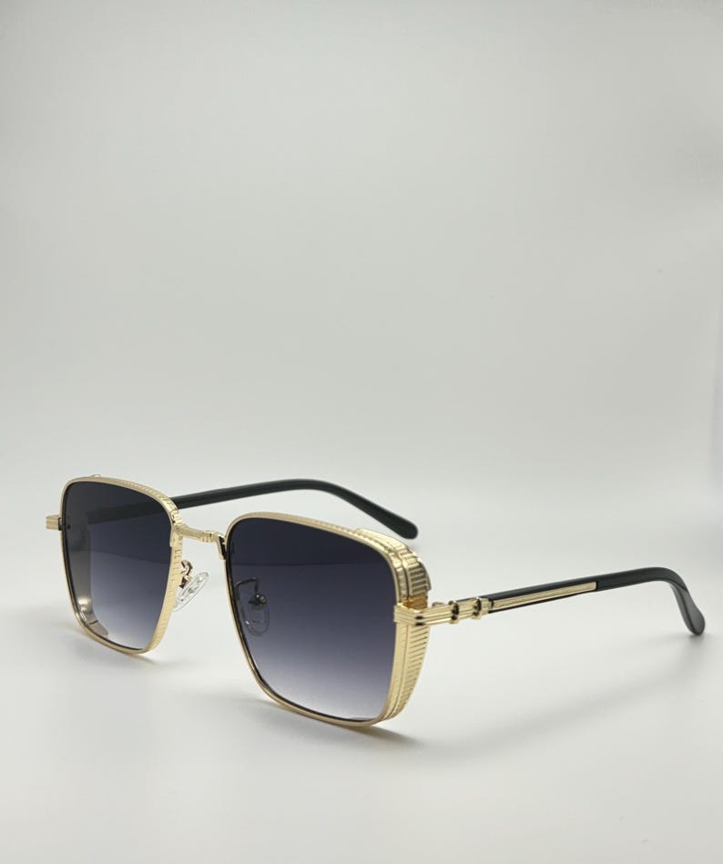Peut inclure: Lunettes de soleil carr&eacute;es avec des montures dor&eacute;es et des verres noirs. Les lunettes ont une monture en m&eacute;tal dor&eacute; textur&eacute;e.