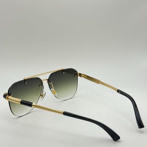 May include: Gold-framed sunglasses with a gradient lens. The sunglasses feature a double bridge and black detailing on the arms. The lenses transition from dark green at the top to a lighter green at the bottom.