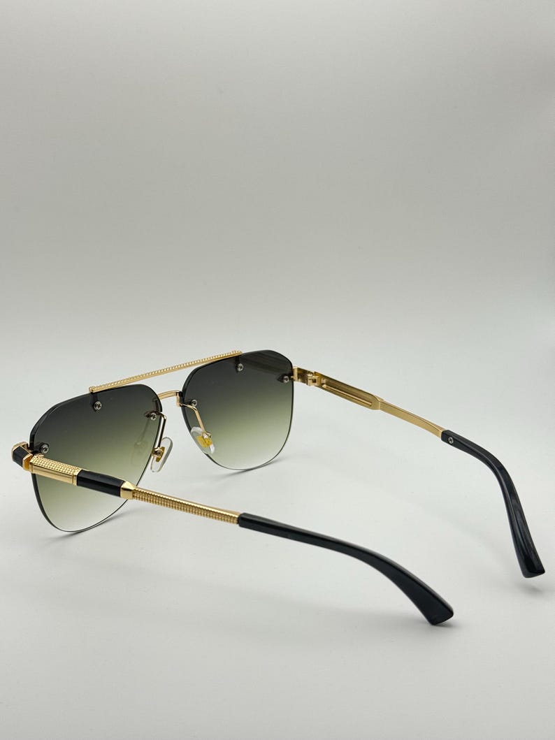 May include: Gold-framed sunglasses with a gradient lens. The sunglasses feature a double bridge and black detailing on the arms. The lenses transition from dark green at the top to a lighter green at the bottom.