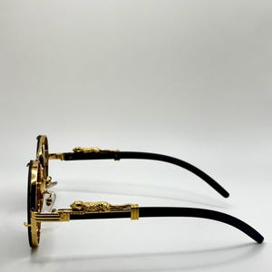 May include: Round sunglasses with gold rims and black arms. The frames have a gold leopard design on the temples. The lenses are dark. The sunglasses are set against a white background.