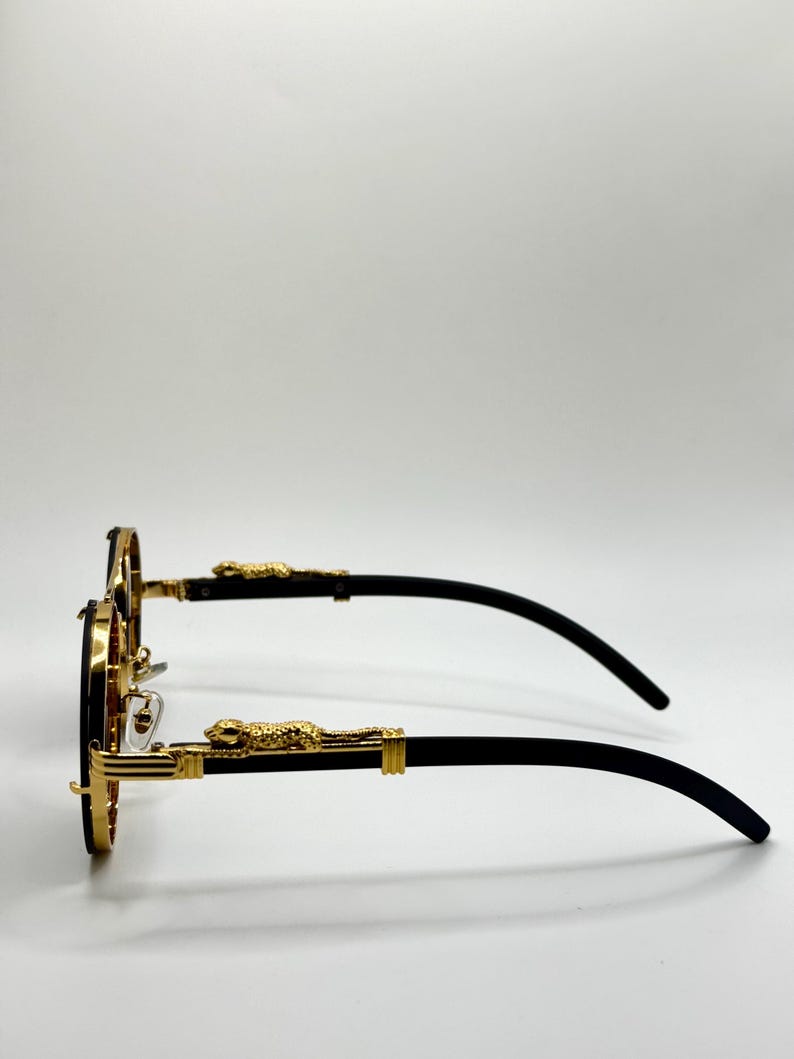 May include: Round sunglasses with gold rims and black arms. The frames have a gold leopard design on the temples. The lenses are dark. The sunglasses are set against a white background.