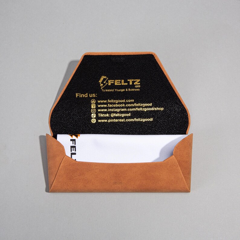 Pode incluir: Porta-cart&otilde;es de couro castanho claro aberto, com interior preto. O porta-cart&otilde;es apresenta o texto "FELTZ GOOD" e informa&ccedil;&otilde;es de m&iacute;dia social em dourado. Um cart&atilde;o de visita branco &eacute; parcialmente vis&iacute;vel no interior.