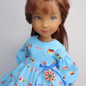 Op de afbeelding: Een blauwe pop met lang bruin haar die een blauwe jurk draagt met daarop gedrukte vlaggen van verschillende landen. De jurk heeft een blauw lint om de taille gebonden met een gouden hanger van een scheepswiel. De jurk heeft het woord "TRAVEL" op de voorkant.
