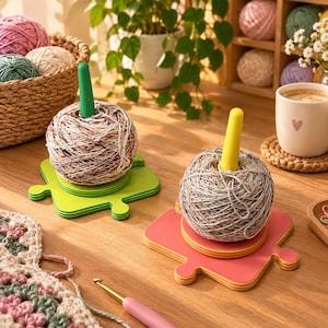 Puede incluir: Dos soportes para ovillos con un eje vertical sobre una base en forma de pieza de puzzle. Uno es verde lima con eje verde y el otro es rosa coral con eje amarillo. Cada uno sostiene un ovillo de lana de colores jaspeados.