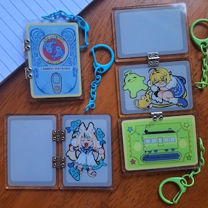 Pode incluir: Uma coleção de chaveiros de acrílico transparente com vários designs. Um chaveiro apresenta um rótulo azul de lata que diz "CANNED MACKEREL". Outros chaveiros têm ilustrações em estilo anime de personagens e um bonde verde. Cada chaveiro tem uma dobradiça de metal e uma corrente.