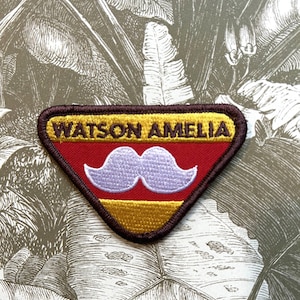 Può includere: Una toppa ricamata a forma di triangolo con un bordo marrone, una striscia inferiore dorata, una striscia centrale rossa e una striscia superiore gialla. La toppa presenta il testo "WATSON AMELIA" in lettere marroni e un baffo bianco.