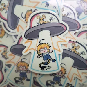 May include: A set of stickers featuring a cartoon character being abducted by a UFO. The character is wearing a blue and white outfit and has yellow hair. The UFO is gray and has a red light on top. The stickers are on a white background.