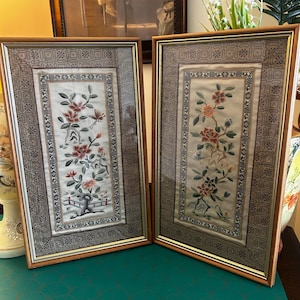May include: Two framed embroidered artworks. Each features a floral design with red and orange blossoms, green leaves, and a small bird. The frames are a warm brown with a decorative border. The background of the embroidery is a light cream colour.