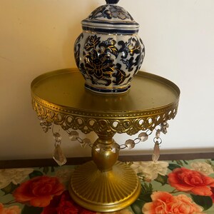 May include: A gold-coloured cake stand with a round top and a decorative, scalloped edge. The stand has dangling crystal accents. A blue and white porcelain jar with a floral design sits on top. The jar has a matching lid.