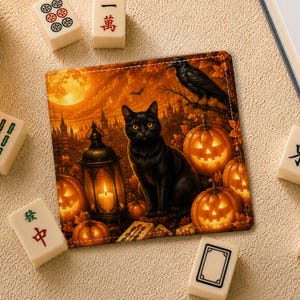 Puede incluir: Un posavasos cuadrado con una escena de Halloween que muestra un gato negro, calabazas brillantes, un farol encendido y un cuervo posado en una rama bajo una luna llena. El fondo muestra la silueta de un castillo en un cielo naranja.