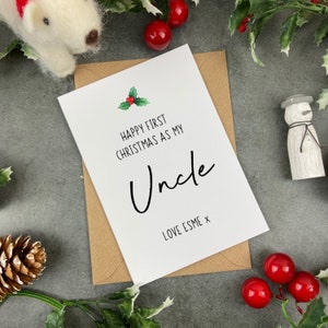 May include: A white card with a green holly sprig and red berries. The card reads "Happy First Christmas as my Uncle" with the word "Uncle" in a black cursive font. The card is signed "Love Esme x".