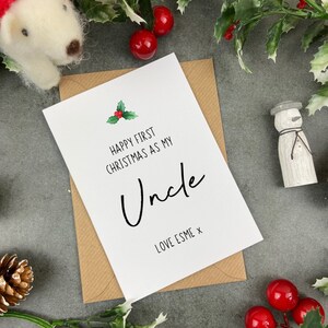 May include: A white card with a green holly sprig and red berries. The card reads "Happy First Christmas as my Uncle" with the word "Uncle" in cursive. The card is signed "Love Esme x".
