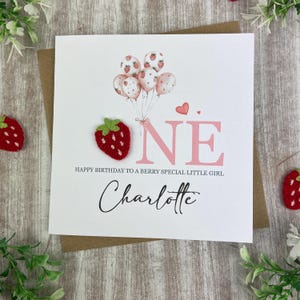 May include: A white birthday card with a strawberry theme. The card features a felt strawberry, pink text that reads "ONE", and a cluster of balloons with strawberry designs. The card also includes the name "Charlotte".