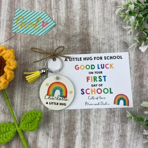 May include: A circular keychain featuring a rainbow design and the name "Charlotte". A card reads "A Little Hug For School, Good Luck on Your First Day of School". A yellow tassel and a "Good" arrow are also present.