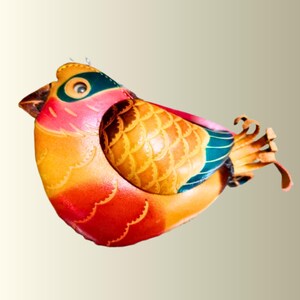 Könnte beinhalten: Ein vogelförmiges Objekt aus mehrfarbigem Leder mit einem lebendigen Farbverlauf in Rot, Orange und Gelb am Körper. Die Flügel zeigen ein strukturiertes, schuppenartiges Muster in Braun- und Goldtönen, akzentuiert durch dunkel-petrolfarbene Flügelspitzen.