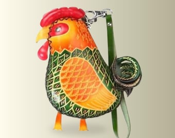 May include: A rooster-shaped leather bag featuring a vibrant orange and yellow head with a red comb and wattle. The body displays a green and yellow scale pattern with a dark green tail. A green leather strap is attached with a silver metal clasp.