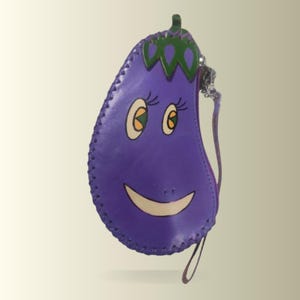 Op de afbeelding: Een paarse, auberginevormige portemonnee met een lachend gezicht. De portemonnee heeft een ritssluiting, een klein polsbandje en groene en witte accenten. De randen zijn gestikt met een contrasterende draad.