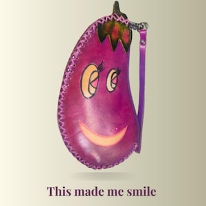 Puede incluir: Una bolsa de cuero morada con forma de berenjena con un diseño de cara sonriente. La bolsa tiene una parte superior verde y marrón, bordes cosidos y una correa morada. El texto "This made me smile" está en la parte inferior.