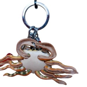 May include: A leather keychain shaped like a crab with a white body and orange and brown legs. The keychain has a silver metal ring.