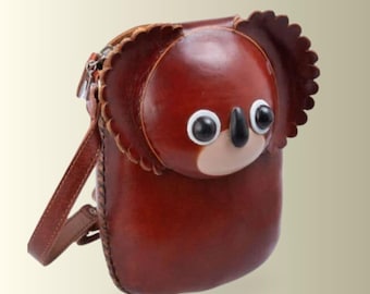 以下が含まれることがあります： コアラの顔の形をした茶色のレザー製ショルダーバッグ。バッグの前面は滑らかで丸みを帯びており、大きな白黒のプラスチック製の目、小さな黒い鼻、スカラップ状の耳のディテールが特徴です。同色の茶色のレザーストラップが付いています。