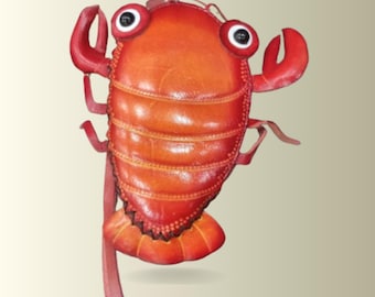 May include: A lobster-shaped bag in shades of orange and red, featuring a segmented body, two raised claws, and two prominent eyes with black pupils. The bag includes a long, thin strap attached to the side, with visible stitching along the edges.