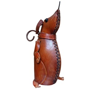 Puede incluir: Una figura de cuero marrón con forma de ratón de pie, con un hocico largo, ojos oscuros y una cola rizada. El cuerpo presenta costuras visibles de color marrón oscuro a lo largo de la costura frontal y un collar decorativo con patrón geométrico.