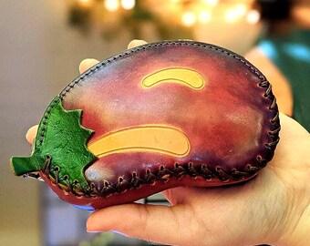 May include: A handcrafted leather coin purse shaped like an eggplant. The purse features a deep purple body with yellow accents and a green leaf detail. The edges are stitched with dark thread.