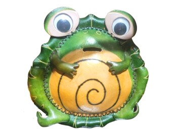 May include: A green and yellow frog-shaped coin bank with large googly eyes. The frog has a dark green outer body with stitched edges and a lighter yellow circular belly featuring a spiral design. A horizontal coin slot is located on the chest.