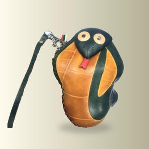 以下が含まれることがあります： コブラの形をしたレザー製のコインケース。頭部と胴体はダークグリーンで、腹部は格子模様のタンカラー、赤い舌が特徴です。2つの丸い目があり、細いレザーストラップには金属製の回転式クラスプが取り付けられています。