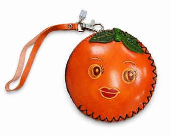 May include: Orange-shaped leather coin purse with a smiling face, green leaf accents, and a wrist strap. The purse has a zipper closure and a silver-tone clasp. The orange-colored purse is round and has a black stitched border.