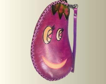May include: A purple leather coin purse shaped like an eggplant with a smiling face. The purse has a stitched border, a small handle, and a wrist strap. The eggplant's stem is green and brown.