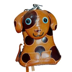 May include: A tan and dark brown spotted dog-shaped leather pouch featuring a metal clasp at the top. The dog has large blue and black eyes, a stitched smile, floppy ears, and a small tail. The surface is smooth with visible dark brown circular spots.