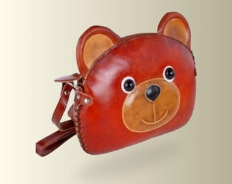 以下が含まれることがあります： クマの顔の形をした茶色のレザー製ショルダーバッグ。2つの丸い耳、2つの黒い円形の目、ダークブラウンの鼻、ステッチされた口元が特徴です。銀色の金属製リベットで取り付けられた、お揃いの茶色のレザーストラップが付いています。