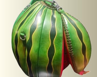 May include: A round, structured handbag shaped like a watermelon, featuring a green exterior with dark green vertical stripes and yellow accents. The bag has a silver-toned metal zipper closure and a bright red interior lining visible at the opening.