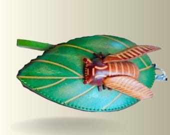 May include: A green leather leaf-shaped coin purse with a brown and tan cicada design. The cicada has detailed wings and is attached to the leaf. The leaf has yellow veins and a green stem.