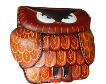 May include: A handcrafted leather owl-shaped bag in shades of brown and orange. The bag features intricate detailing, including stitched patterns and decorative elements. The owl's eyes are white with black accents, and the bag has a flap closure.