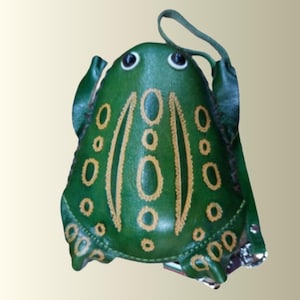 May include: A green frog-shaped coin purse featuring a smooth surface with embossed yellow oval patterns, two dark eyes, and a thin green strap attached to the side. The design mimics the form of a frog with defined limbs and a rounded body shape.