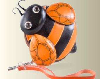 May include: A coin purse shaped like a bee with a black and orange striped body, two orange wings with a geometric pattern, a black head with two eyes, and two antennae. An orange leather wrist strap is attached to the side with a metal clasp.