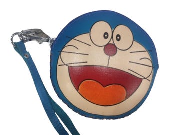 May include: A blue, round coin purse featuring the cartoon character Doraemon. The purse has a blue wrist strap and a silver clasp. The character's face is cream-colored with a red mouth and orange tongue. The purse is a fun accessory.