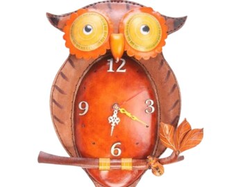 May include: A wall clock shaped like an owl with brown leather-like texture, large orange-rimmed eyes, and a central oval face featuring white numerals 3, 6, 9, and 12. The owl perches on a brown branch with three leaves and has ornate clock hands.