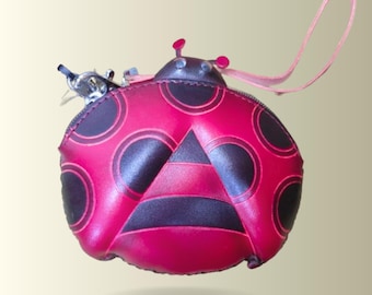 May include: A red ladybug-shaped coin purse with black spots and a black and red striped design. The purse has a zipper closure, a small pink strap, and a silver key ring. The ladybug's head has two small red antennae.