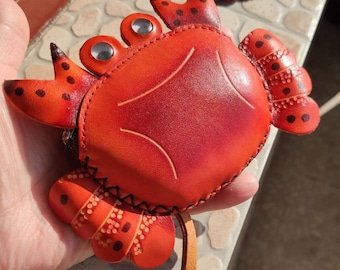 May include: A red leather coin purse shaped like a crab with a smooth, rounded body, two raised eyes with silver centers, and articulated legs featuring dark brown spots. The edges are stitched with black thread, and a tan leather pull tab is attached.