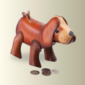 May include: A handcrafted leather dog-shaped coin bank featuring a rich brown body with a lighter tan face, dark stitched edges, and a small tail. The figure has black eyes and a black nose, with a coin slot on the back and three metal coins nearby.
