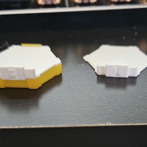 May include: Three hexagonal game pieces, two are white with a raised number on each, one is white with a yellow base and a raised number on each.