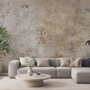 Könnte beinhalten: Ein hellgraues Ecksofa mit mehreren Zierkissen, einem runden beigen Couchtisch und einer Topfpalme. Die Wand hinter dem Sofa zeigt eine strukturierte, verwitterte Oberfläche in Braun-, Grau- und Goldtönen, die an gealterten Putz erinnert.