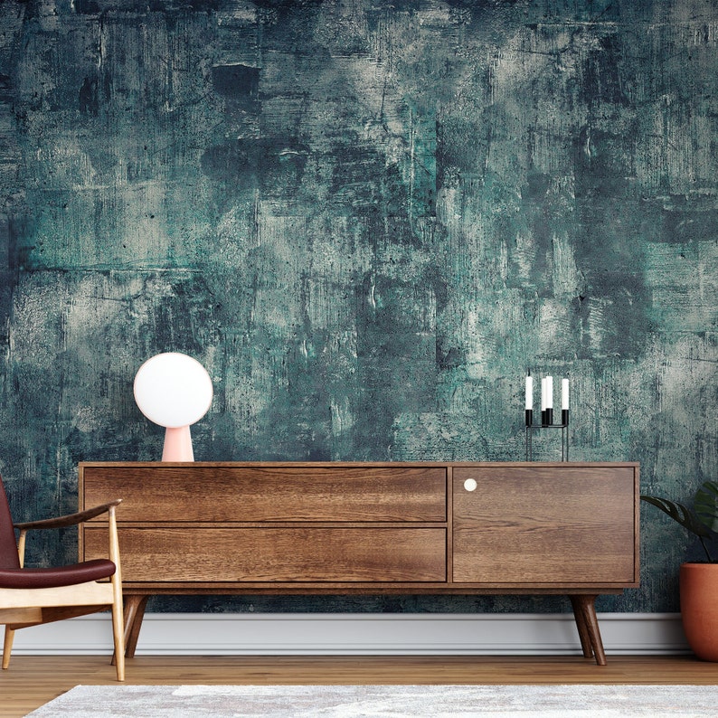 K&ouml;nnte beinhalten: Ein Sideboard aus Holz im Mid-Century-Modern-Stil mit zwei Schubladen und einer Schrankt&uuml;r. Eine rosa Tischlampe mit wei&szlig;em Kugelschirm steht darauf. Der Hintergrund ist eine t&uuml;rkisfarbene und graue, abstrakt strukturierte Wand.