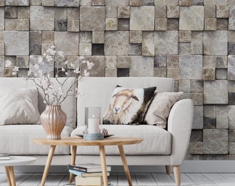 May include: A white sofa with pillows sits in front of a wall covered in square, brown and gray textured tiles. A wooden coffee table with a stack of books sits in front of the sofa. A vase with white flowers sits on the table.