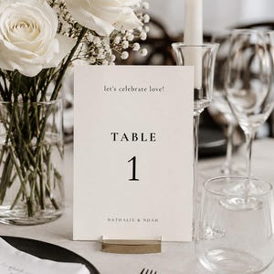 Könnte beinhalten: Eine rechteckige, cremeweiße Tischnummernkarte, die von einem goldfarbenen Metallständer gehalten wird. Die Karte zeigt den Text let's celebrate love!, TABLE, 1 und NATHALIE & NOAH in einer schwarzen Serifenschrift. Sie steht aufrecht.