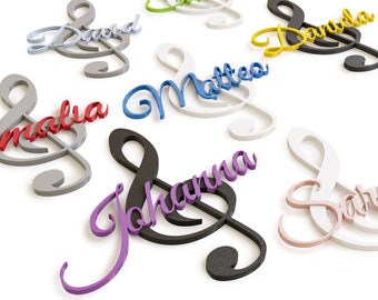 May include: A collection of personalised wooden treble clef ornaments in various colours, including red, blue, purple, black, white, and yellow. Each ornament features a name written in a cursive font, such as "Malia", "Matteo", "Johanna", "David", "Daniela", and "Sara".