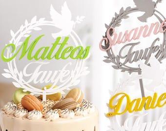 May include: Two white cake toppers with a wreath design and a dove. The toppers have the names "Matteo Taufe" and "Susanne Taufe" written in green and pink letters. A third topper has the name "Daniel Taufe" written in yellow letters.